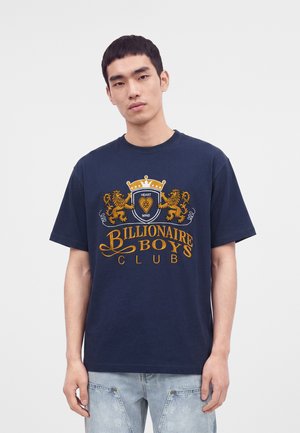 Joven con camiseta azul marino de Billionaire Boys Club con escudo dorado y vaqueros azul claro, de pie frente a un fondo liso y claro.