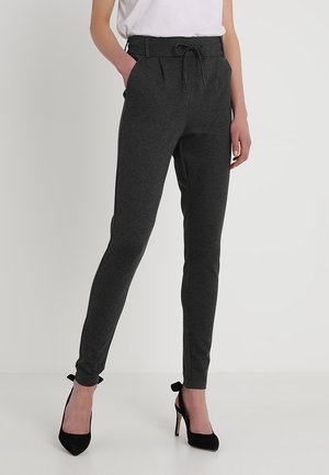 Kvinde iført mørkegrå slim-fit bukser med bundet taljebånd og sorte højhælede sko, stående mod en ensfarvet lys baggrund.
