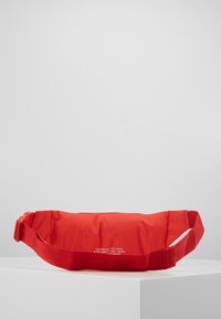 Borsa in nylon rossa a vita con superficie texturizzata, cinturino regolabile e zip. Testo visualizzato sul davanti in bianco. Design elegante e compatto.