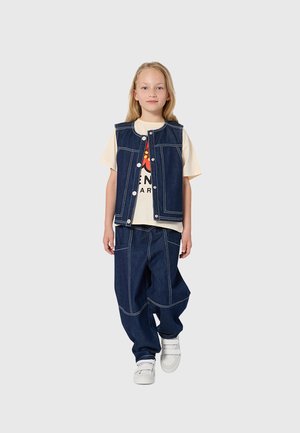 Jong meisje dat loopt, gekleed in een licht T-shirt met grafische print, een donkerblauw denimvest en een broek met witte stiksels, en witte sportschoenen.