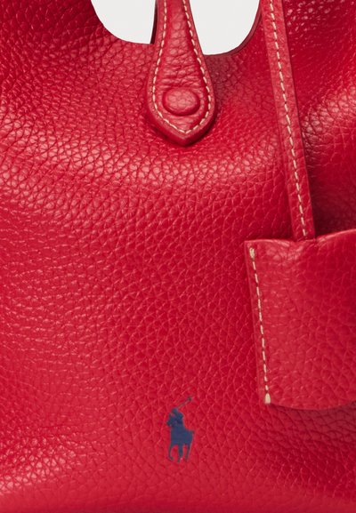 Sac à main en cuir rouge avec une texture grainée, présentant un sommet arrondi, une fermeture à bouton et une petite poche extérieure. Écusson de logo bleu visible.