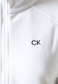 Bílá fleecová mikina na zip s jemnou texturou, vyšité logo "CK" v černé barvě a kontrastním zapínáním na zip.