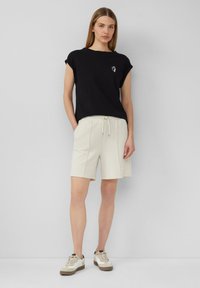 Zwarte katoenen T-shirt met opgerolde mouwen en een klein wit logo, gecombineerd met crèmekleurige shorts en lichte sneakers met textuuraccenten.