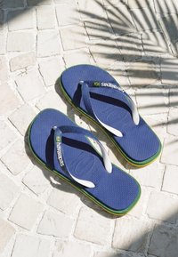 Μπλε σαγιονάρες Havaianas με λευκά λουριά και λεπτομέρεια της σημαίας της Βραζιλίας σε λευκό πλακάκι δάπεδο με σκιά από φοίνικα.