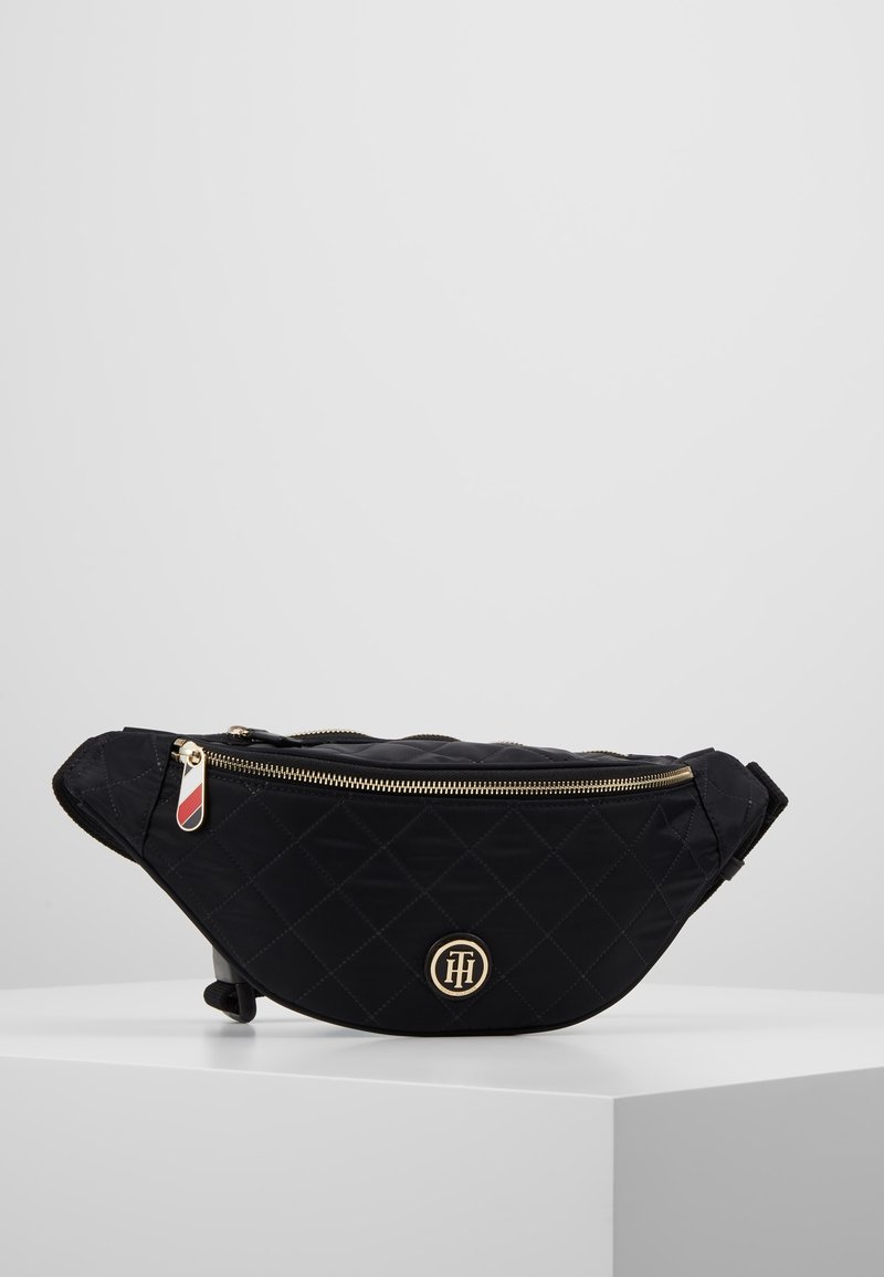 Sac banane noir matelassé avec fermeture éclair dorée et emblème du logo, présenté sur une surface blanche sur fond gris uni.