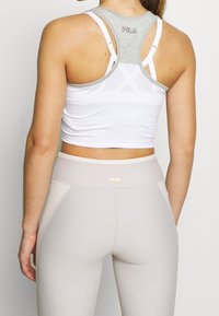 Bílý crop top se šedou zadní částí a ramínky, v kombinaci se světle šedými legínami s vysokým pasem a malým zlatým logem.