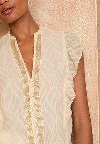 Light beige blouse with a textured wavy pattern, a gold sequined front panel, ruffled sleeves, and button fastenings.