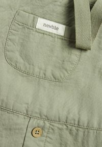 Vêtement en tissu vert clair avec une poche plaquée comportant une étiquette indiquant "newbie" et un détail de bouton beige. Design texturé et décontracté.