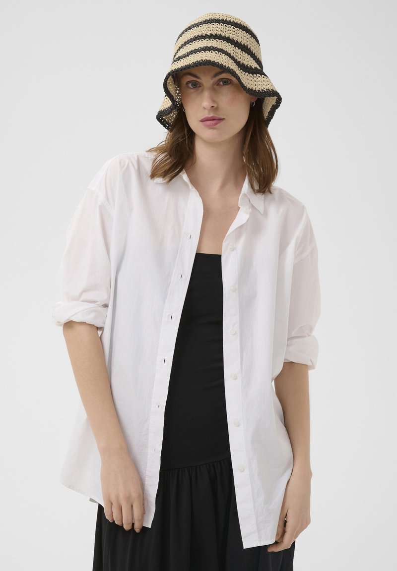 Femme portant une robe noire, une chemise blanche oversize à boutons avec manches retroussées, et un chapeau seau en crochet beige et noir rayé.