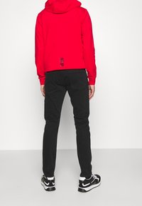 Červená mikina s kapucí a přední kapsou ve stylu klokan, černé slim-fit džíny a černé tenisky s bílými akcenty. Výrazný detail na zadní straně mikiny.