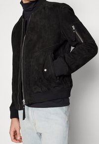Giacca bomber in suede nera con polsini e orlo a coste, dotata di una tasca laterale con zip e zip frontale. Texture liscia con dettagli di design minimal.
