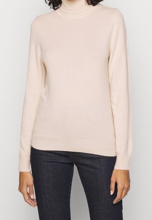 Kvinde iført en beige rullekravetrøje og mørkeblå jeans, stående mod en ensartet lys baggrund.