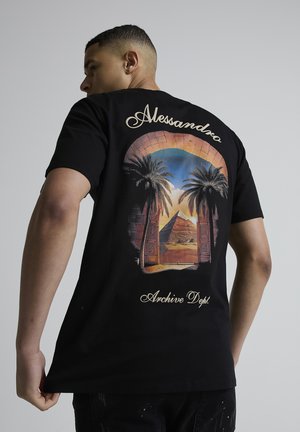 Mand iført sort t-shirt med ørkenlandskab, palmer og pyramidegrafik indrammet af en bue, med teksten "Alessandro" og "Archive Dept.".