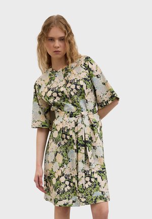 Frau mit welligen blonden Haaren, die ein knielanges Blumenmusterkleid mit grünen, blauen und beigen Mustern sowie einem Stoffgürtel in der Taille trägt.