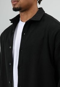 Zwarte fleece blouse met knopen en een kraag, met zwarte knopen en een wit ondershirt, met een textuur van de stof zichtbaar.