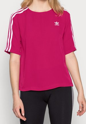 Kobieta w różowej koszulce Adidas z białymi paskami na rękawach i czarnych spodniach z białymi paskami po bokach.