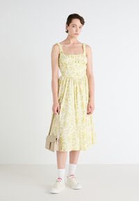 Žlté kvetinové šaty so štvorcovým výstrihom, nariaseným pásom a plisovanou sukňou. Kombinované s bielymi teniskami a malou béžovou kabelkou.