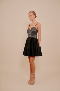 Žena u bezrukavoj crnoj haljini s vrhom od šljokica i slojevitom suknjom, nosi zlatne okrugle naušnice i srebrne sandale s trakama.