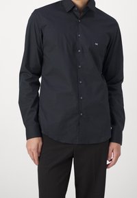 Camicia button-up blu scuro con maniche lunghe, leggera lucentezza e un piccolo logo bianco sul lato sinistro del petto. Texture liscia, orlo arrotondato.
