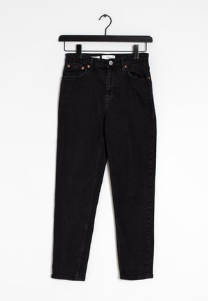 Jean noir taille haute suspendu sur un cintre en bois noir contre un fond blanc, avec des poches avant et une fermeture à bouton.