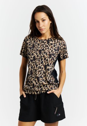 Vrouw staat met handen in haar zakken, draagt een kort mouwshirt met luipaardprint en zwarte shorts tegen een effen witte achtergrond.