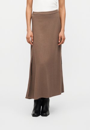 Brauner langer Rock mit Seitenschlitzen, getragen mit schwarzen Knöchelstiefeln und einem weißen ärmellosen Oberteil, dargestellt von der Mitte des Torsos bis zu den Füßen.