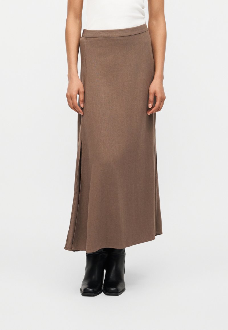 Καφέ μακριά φούστα με πλευρικές πλαϊνές σκισίματα, φορεμένη με μαύρες μποτάκια αστραγάλου και λευκό αμάνικο τοπ, φαίνεται από τη μέση μέχρι τα πόδια.