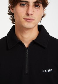 Pull en polaire zippé noir avec col, arborant un petit logo blanc "VOLCOM" sur le côté gauche et une texture lisse et douce.