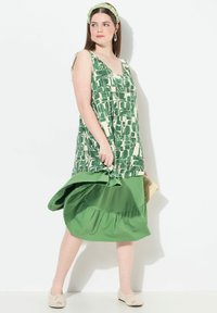 Vestido sin mangas de estampado verde y crema con una falda verde en capas, combinado con una diadema verde y un bolso de mimbre, lucido por una modelo.