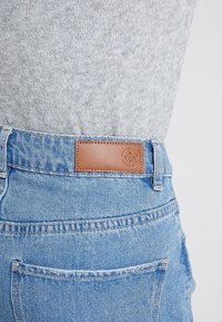 Ljusblå denimjeans med en brun läderlapp på baksidan av midjan. Funktioner inkluderar fransig fåll och standard bakfickor.