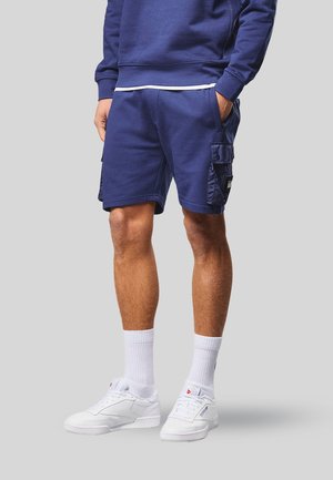 Hombre vistiendo pantalones cortos cargo azul marino, sudadera a juego, calcetines blancos hasta el tobillo y zapatillas blancas, de pie frente a un fondo liso.