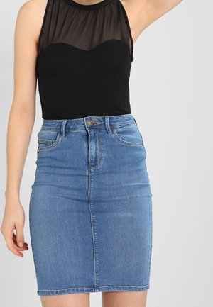 Kobieta w czarnym topie bez rękawów z prześwitującą górną częścią i wysokim stanem, niebieską spódnicą ołówkową z denimu, stojąca na jednolitym tle.