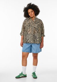 Blouse à imprimé zèbre avec coupe ample, design boutonné en beige et noir, associée à un short en denim. Des baskets vertes et des chaussettes blanches complètent le look.