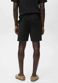 Homme debout, portant un short noir texturé et un t-shirt noir assorti, associé à des chaussures à enfiler marron clair, de dos sur fond uni.
