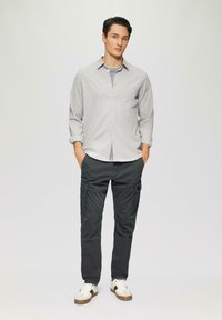 Lichtgrijze button-up shirt, donkere cargo broek met zakken, en witte sneakers met bruine accenten. Model staat tegen een neutrale achtergrond.