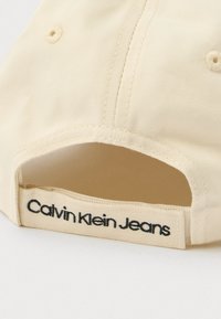 Krems krāsas beisbola cepure ar izliekto vizieri, izgatavota no teksturēta auduma, aizmugurē ar melnu izšūtu "Calvin Klein Jeans" etiķeti.