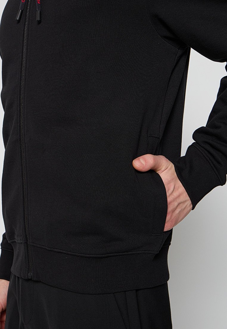 Eine Person trägt eine schwarze Reißverschlussjacke, mit einer Hand in die seitliche Tasche der Jacke gesteckt.