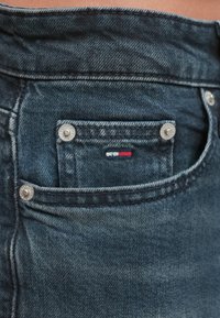 Σκούρο μπλε τζιν με μικρή μπροστινή τσέπη, ασημένια μεταλλικά στοιχεία και κεντημένο λογότυπο σε κόκκινο, λ�ευκό και μπλε στην τσέπη.