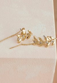 Zlati uhani z motivom lista, ki obsega tri liste in verigo, okrašeni s prozornimi gemi v središču, ležijo na teksturirani površini.