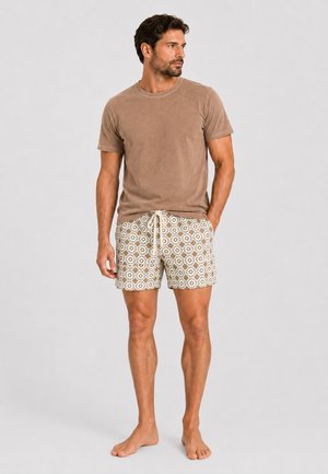 Mann steht barfuß, trägt ein braunes kurzärmeliges Hemd und beige gemusterte Shorts mit Kordelzug, schaut nach links.