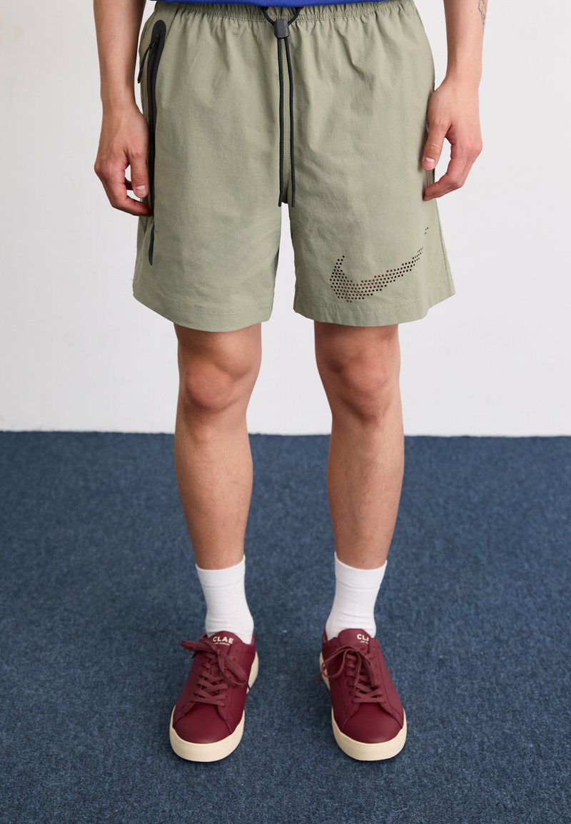 Shorts Nike en vert clair, fabriqués en tissu léger. Les caractéristiques incluent des accents noirs, une taille ajustable avec cordon de serrage, et un logo perforé sur le côté.