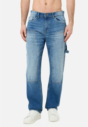 Mann trägt zweifarbige blaue Jeans mit großen Kniepatches vorne, seitlichem Schlaufen-Detail und weißen Sneakers, steht vor weißem Hintergrund.