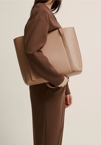 Beige ToteBag mit einem strukturierten Design, glatter Textur und zwei Griffen. An der Seite zu einem braunen Outfit getragen, minimale Markenkennzeichnung sichtbar.