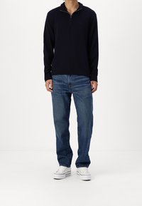 Pullover blu navy con zip al collo, maniche lunghe, polsini a coste, abbinato a jeans in denim blu, vestibilità comoda e sneakers bianche su uno sfondo neutro.
