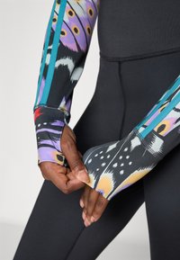 Mâini care trag de mânecile colorate cu model de fluturi, prevăzute cu găuri pentru degete, peste colanții negri și ajustați.