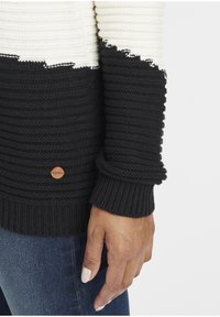 Schwarzer und cremfarbener gestrickter Pullover mit gerippter Textur und rohem Saum. Verfügt über ein rundes Holzlogo-Anhänger an der unteren linken Seite.