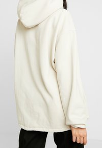 Miękka, oversize'owa bluza z kapturem w kolorze ecru, z regulowanym sznurkiem w kapturze, opuszczanymi ramionami i mankietami na rękawach. Prosty detal przeszycia u dołu.