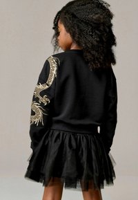 Sudadera negra con bordado de dragón dorado en las mangas, combinada con una falda de tul negra de capas. Tejido suave con una textura blanda.