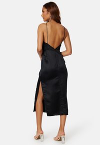 Abito midi in satin nero con spalline sottili, scollatura profonda sulla schiena, apertura laterale e texture liscia. Presenta una silhouette aderente e una leggera lucentezza.