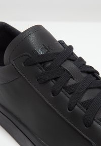 Detailní záběr na černou koženou tenisku s černými šňůrkami, ukazující šité okraje a embosované logo značky na jazyku.